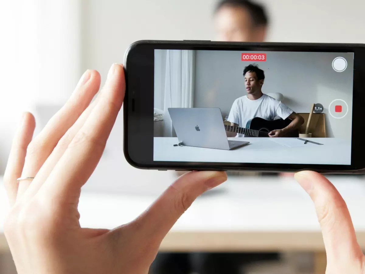 A person is holding a smartphone and taking a video of another person working on a laptop.